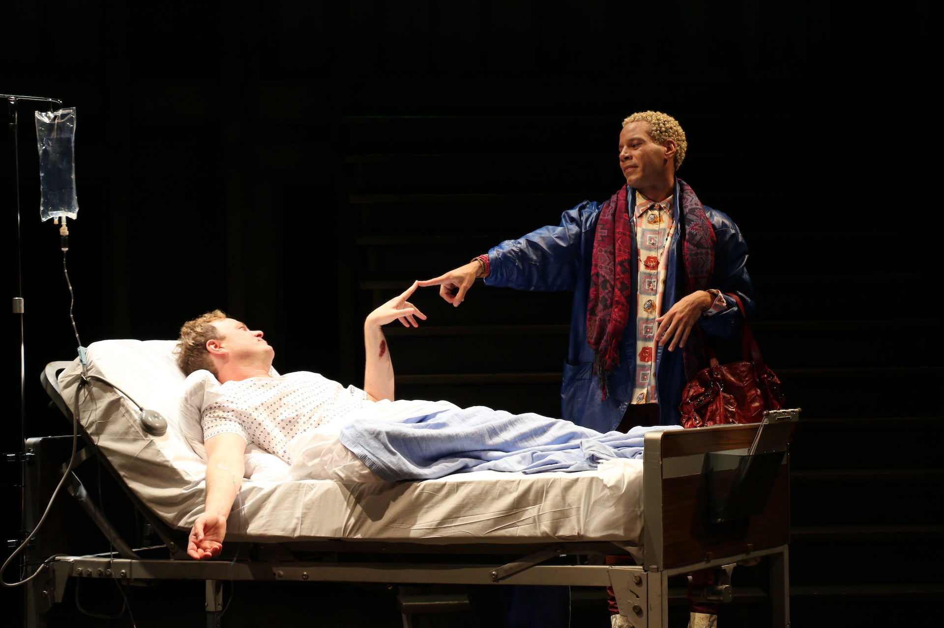 Two actors of the play Angels in America Part I: Millennium Approaches. One of them is in a hospital bed, the other stands next to him, their fingers are touching