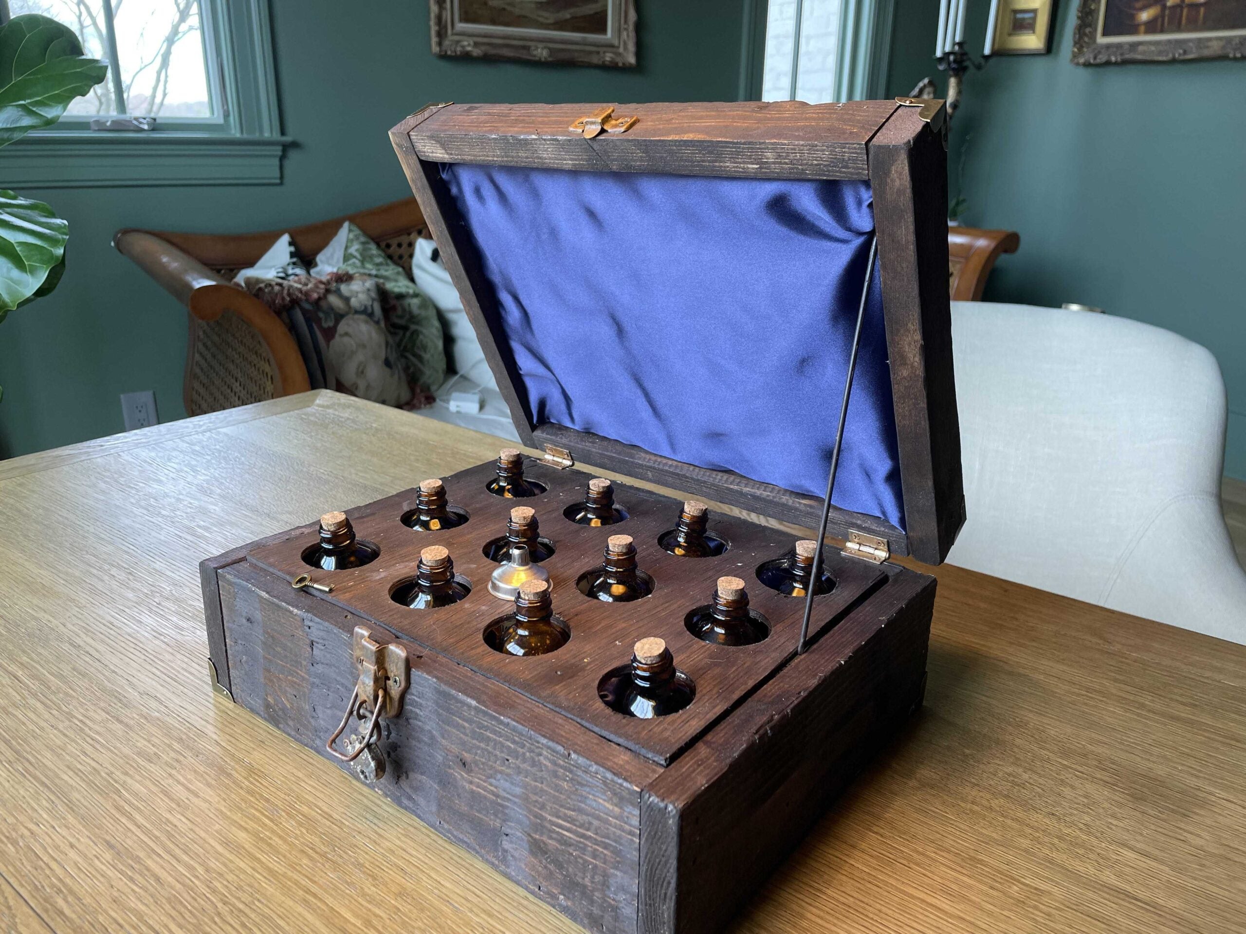 Recreation of an apothecary crate (open), showing a dozen small bottles inside. The top lid is covered by a blue fabric. The crate is on top of a table. There are a sofa and a chair on the background