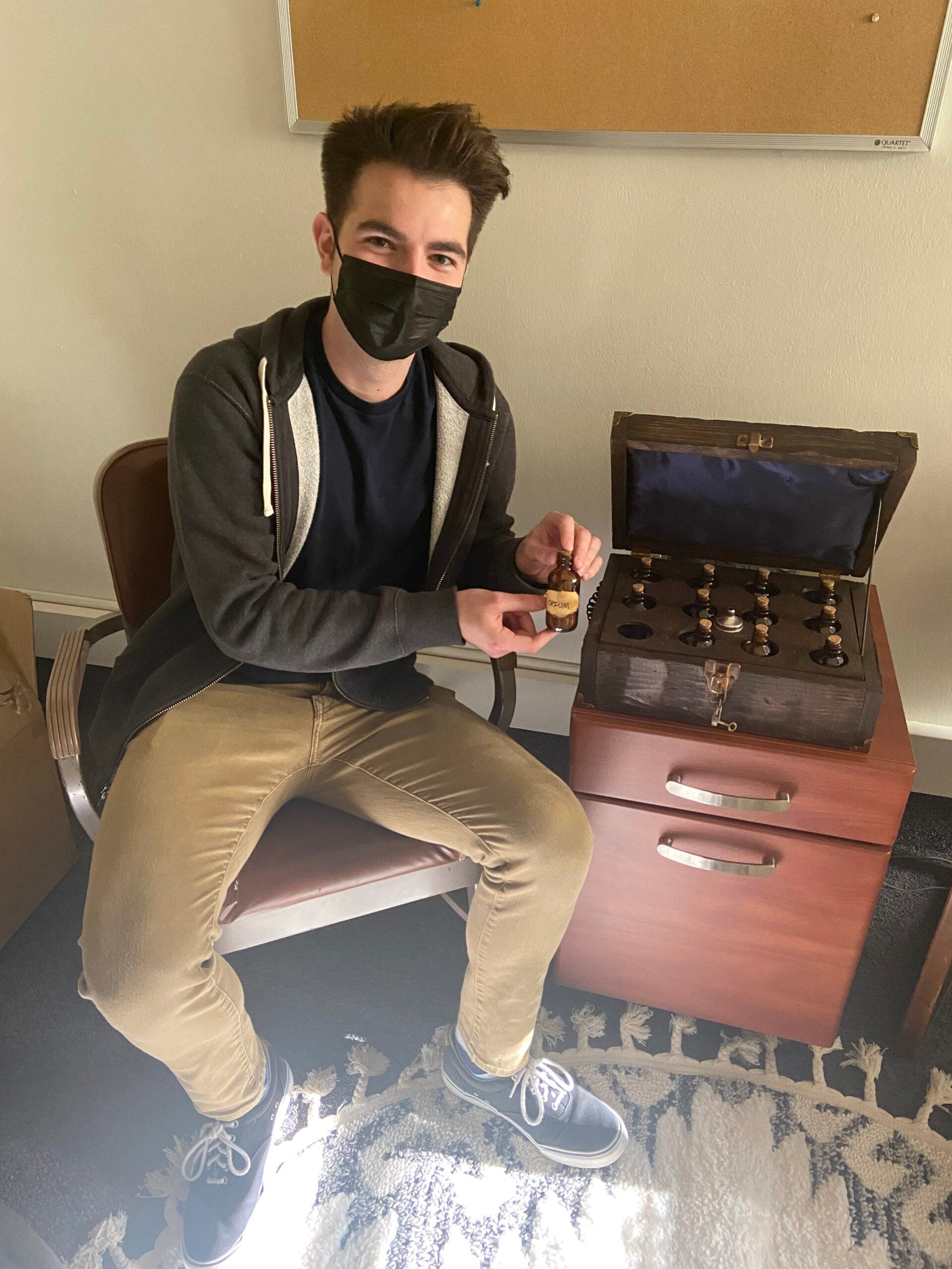 Evan Navori sitting next to the 1820s apothecary crate he recreated, which is open. Evan is holding a small bottle, inside the crate there are 11 more bottles. The crate is on top of a drawer.