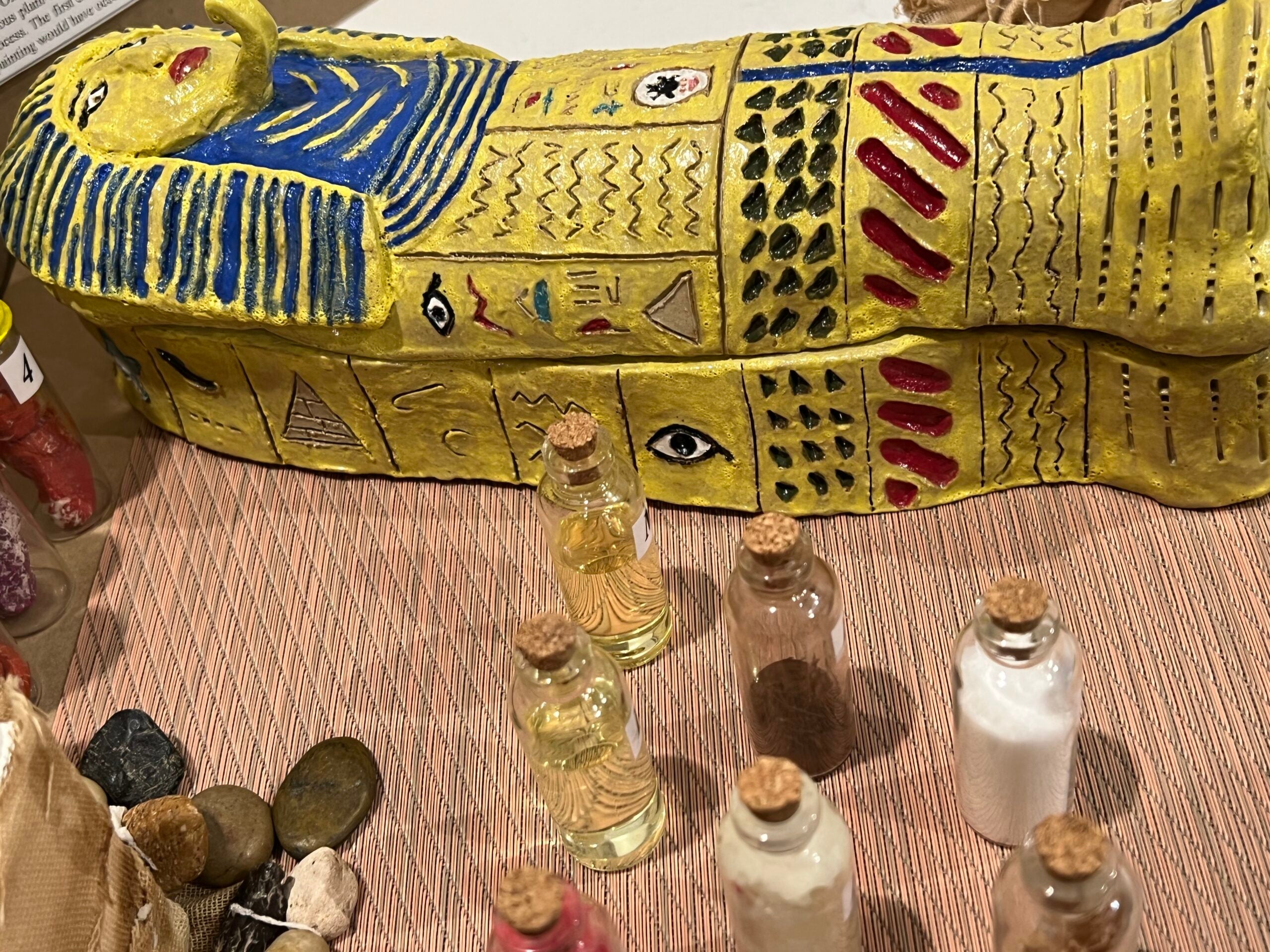 Hand-made clay sarcophagus displaying the final wrapped version of the mummy (the lid is closed). Next, there are bottles representing the plant-based materials used in the mummification process