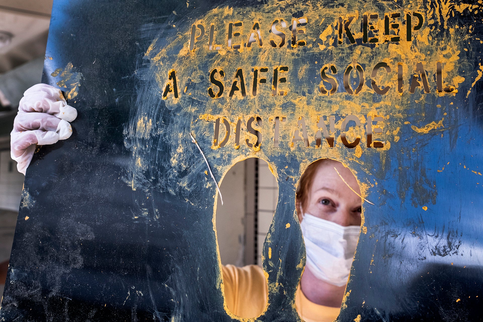 Person wearing a mask, holding a piece of metal with the words "Please keep a safe social distance"