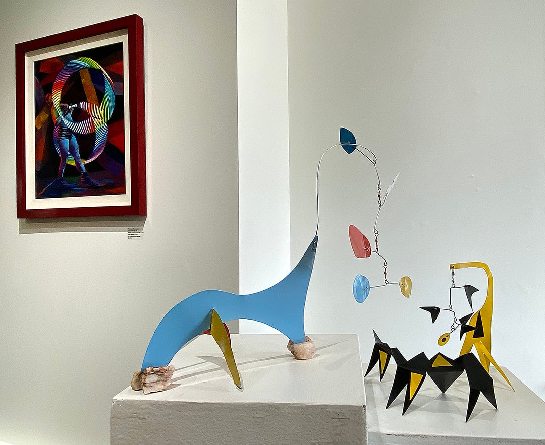 Tony Lee Sangastiano's work on display at the Studio Place Arts. Her work, on the left, shows a circus artist holding giant hula hoops, which, framed in the background, surround the artist entirely. The shapes's have the colors of the rainbow. On the right, there's a sculpture of geometrical figures resembling animals.