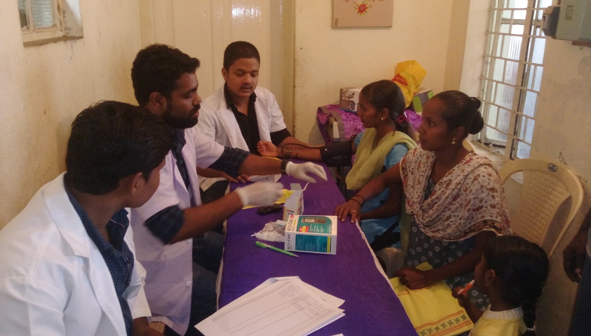 Two Indian women and a child on diabetes consultation, with three medical personnel on a counter, with forms and a device for measuring the levels of sugar on the blood