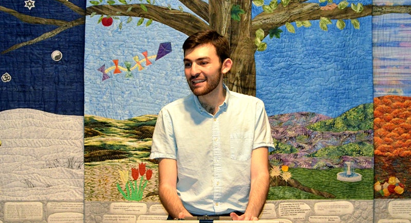 Benjamin Hack smiling. There are three tapestries in the background, representing a landscape with a tree with a kite at day and night, in spring and fall.