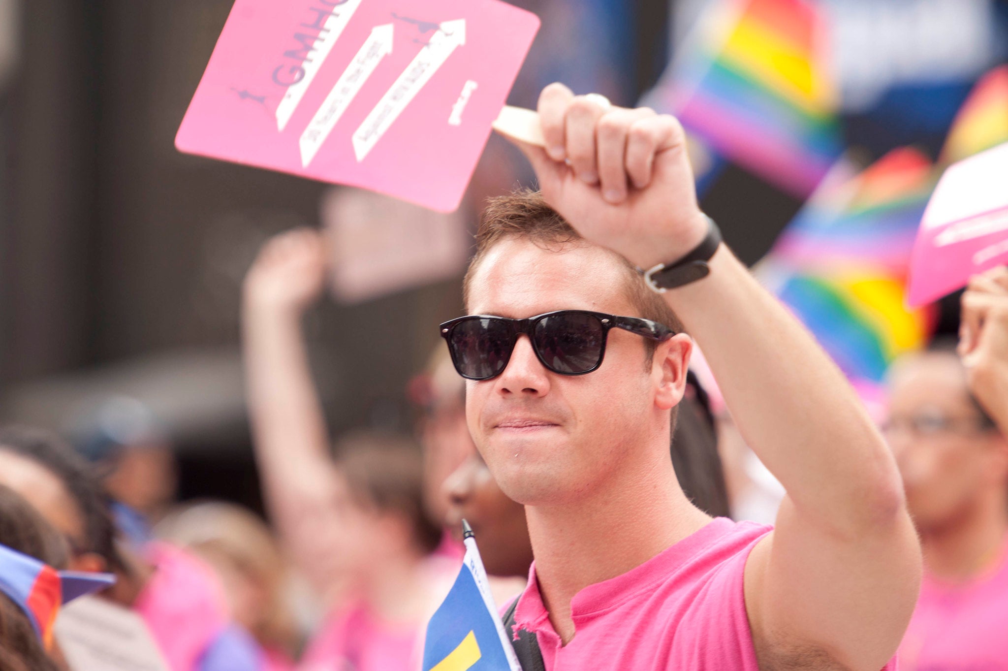 Man wearing sunglasses, a pink t-shirt and holding a pink sign. On the background, numberous rainbow flags