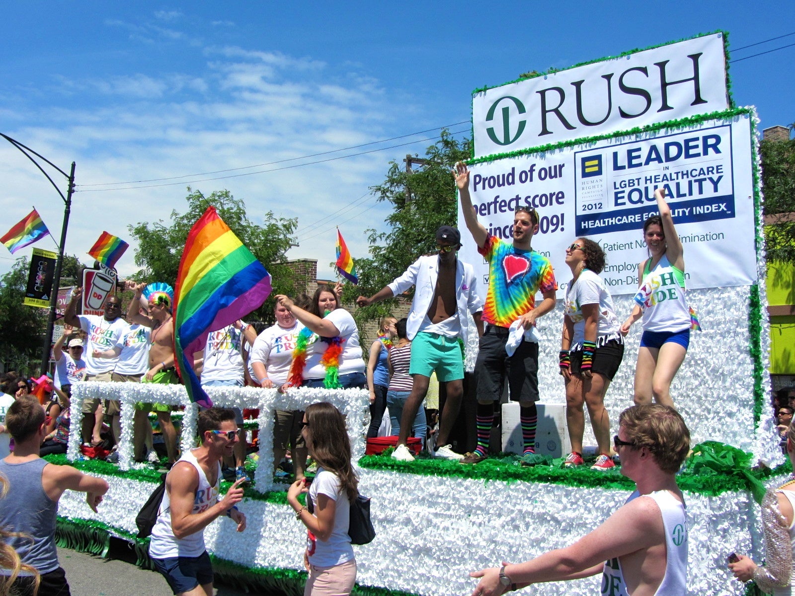 White float on pride parade with young people waving rainbow flags and the logo of the Rush University Hospital and signs that say: 