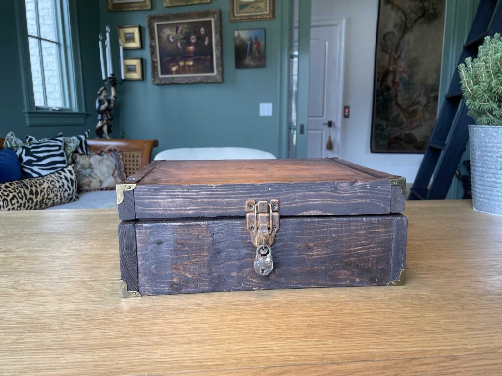 Recreation of an apothecary crate on top of a table (closed). There is a plant next to it, and a sofa, door and some paintings on the background