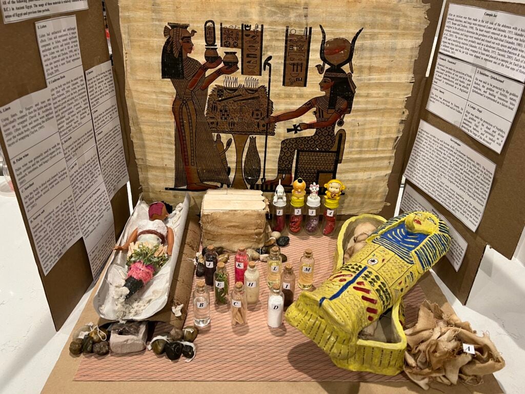 Picture of Layan Shahrour's research project. On the left, there is a doll representing the body that would be wrapped as a mummy in a sarcophagus. On the center, there are bottles representing the plant-based materials used in the mummification process, as well as canopic jars that were used to house their respective organs in line with the figures' spiritual significance and protective power. On the right, there is a hand-made clay sarcophagus displaying the final wrapped version of the mummy (the lid is open). On top, there is a painting showing the human or godly figures associated with death in the New Kingdom of Ancient Egypt. There is also some illegible text on each side, describing the research project