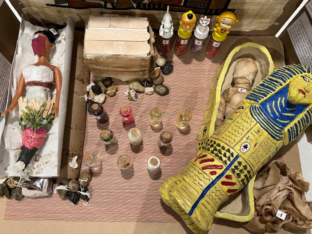 Picture of Layan Shahrour's research project. On the left, there is a doll representing the body that would be wrapped as a mummy in a sarcophagus. On the center, there are bottles representing the plant-based materials used in the mummification process, as well as canopic jars that were used to house their respective organs in line with the figures' spiritual significance and protective power. On the right, there is a hand-made clay sarcophagus displaying the final wrapped version of the mummy (the lid is open)