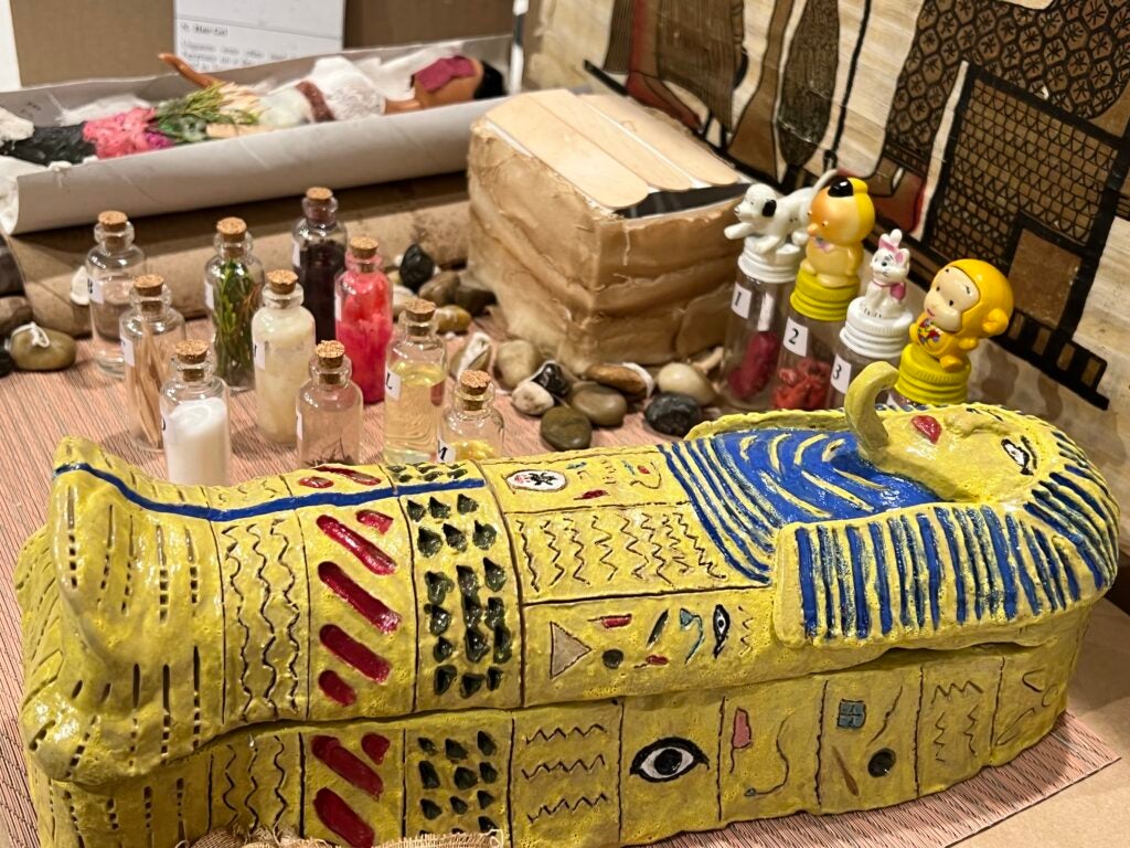 Hand-made clay sarcophagus displaying the final wrapped version of the mummy (the lid is closed). Next, there are bottles representing the plant-based materials used in the mummification process. There are also canopic jars that were used to house their respective organs in line with the figures' spiritual significance and protective power, and a doll representing the body that would be wrapped as a mummy in a sarcophagus